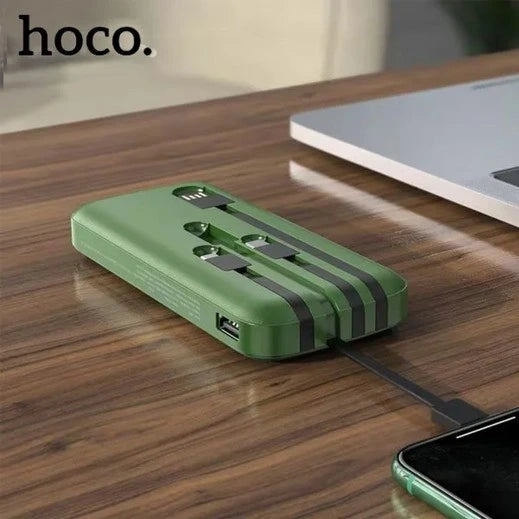 A lifestyle image of the green Hoco DB07A 20000mAh high-capacity power bank from LebTech Store Lebanon, shown on a wooden desk. The power bank is lying flat with its four built-in charging cables (USB, Type-C, Lightning, and Micro-USB) neatly stored in their integrated slots, while one cable is extended to charge a smartphone.
