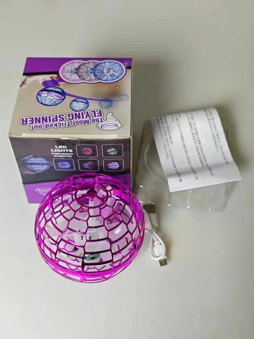 An unboxed pinkish purple flying spinner ball next to its original product packaging box, white USB charging cable, and user manual from LebTech Store Lebanon.