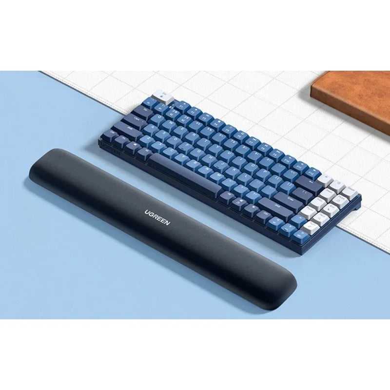 A black Ugreen memory foam keyboard wrist rest paired with a blue and white mechanical keyboard on a desk, available at LebTech Store Lebanon. The image shows the product in a real-world setup, demonstrating its size and sleek ergonomic design.
